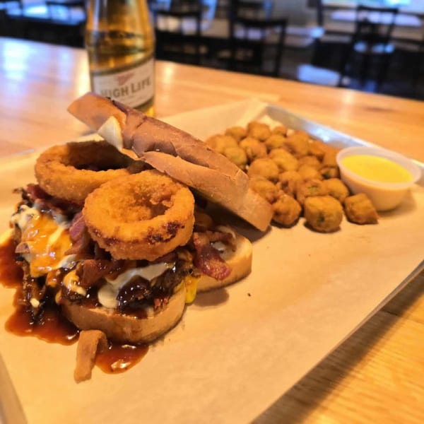 A BBQ burger with onion rings, fried okra, and a Miller High Life beer on a wooden table.