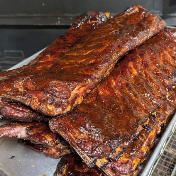 Close-up of several racks of glistening, dark brown BBQ ribs on a metal tray.